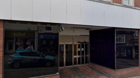 An empty shop. The windows are blacked out and there are two sets of doors in the middle, one of which is automatic. A lone CCTV camera points down towards the entrance.