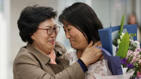 Two women, one older and one younger, embrace. They look like they are crying with Joy, and one is also holding a bunch of flowers