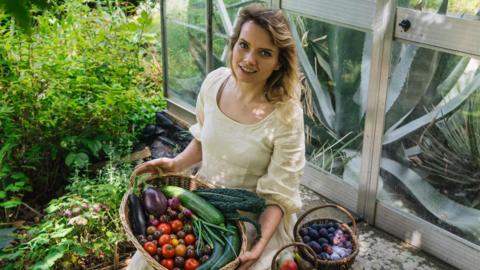 A young woman with brown hair is wearing a flowy cream dress. She is smiling and is sitting by the edge of a greenhouse with a large wicker basket filled with fresh vegetables on her lap. In the background is a lush green garden.