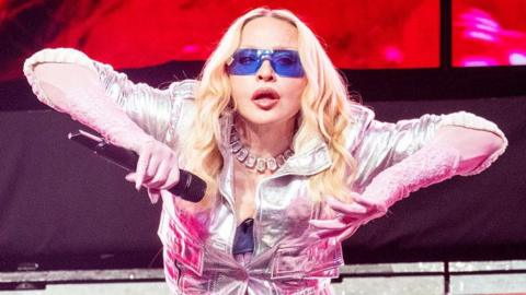 Madonna on stage in New York. She's holding her arms to the side, revealing pink lace gloves beneath a silver jacket. Her eyes are hidden behind jet blue sunglasses, but she appears to be staring directly at the camera.