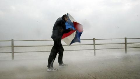 A man walking along a promenade, struggling against the wind. He is holding an umbrella in front of his face, using it as a shield against the wind. The red, white and blue umbrella is only half open. There is mist from the spray from the sea and rain around the man. He is wearing a black coat and black and grey tracksuit bottoms and trainers. 