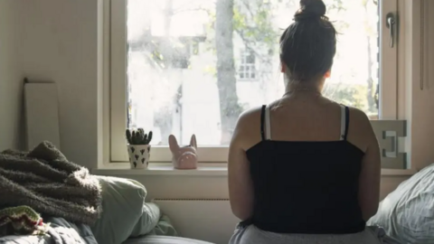 A young woman looks out of a w window with her back to the camera. She is wearing a vest top and is sat in a bedroom.