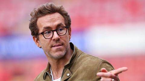 Ryan Reynolds, wearing glasses and a brown jacket, pointing a finger