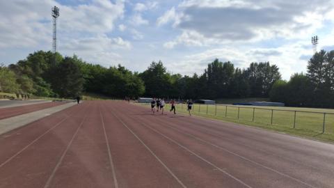 A group of runners on an athletics track sprint on the inside lanes next to a grass field