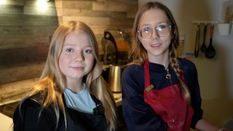 Two young people, Molly and Sinead, are pictured in a modern kitchen setting, standing side by side near a counter top. Both are wearing aprons. Molly on the left has long blonde hair, a light blue T-shirt and a black apron. She is smiling. Sinead has long brown hair, one length in a plait, and is wearing glasses and a red apron. Behind them, there are stainless steel appliances and utensils hanging in a contemporary-looking cooking psace.
