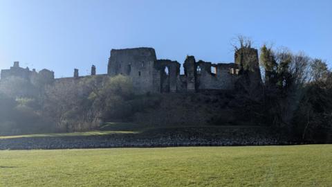 A general view of Cockermouth Castle from the footpath along the River Derwent. The large structure has missing windows and appears to be like a ruin on the right hand-side, while it looks more like a complete structure on the left. The castle overlooks the river and is in a slightly raised position from the riverbank.
