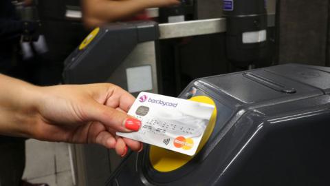 A Barclaycard credit card being placed on a contactless card reader at a train station ticket barrier.