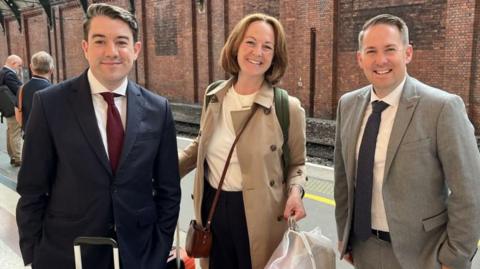 The three new MPs stand on a platform at a train station smiling at the camera