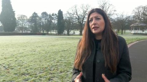 Preet Gill, MP, pictured in a park with long dark hair over her shoulders. She is wearing a dark grey buttoned up jacket.