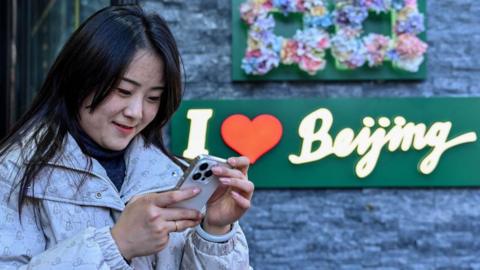 A Chinese woman checks her phone, with a sign in the background reads "I heart Beijing"