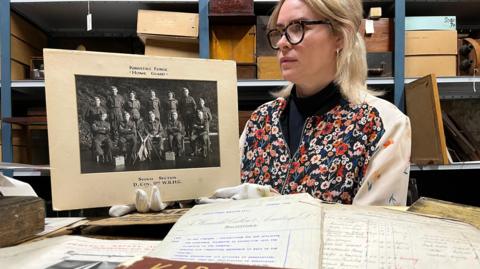 A woman with blonde hair and glasses holds a black and white photograph wearing white gloves. In front of her are a selection of handwritten documents