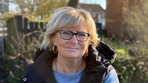 Sue Mitchell smiles at the camera. She has short blonde hair and wears glasses, a black coat and a blue top underneath. She also wears gold hoop earrings.