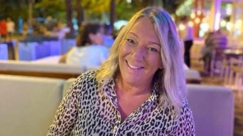 A blond woman wearing a leopard-print blouse sits on a booth. She has her head slightly tilted to the right and smiles directly at the camera.