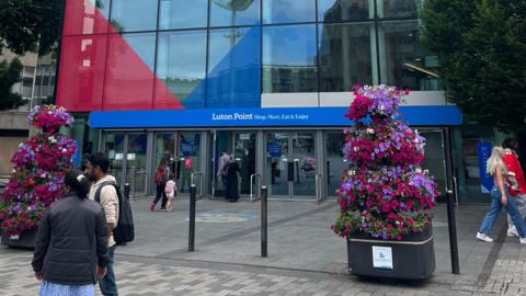 The entrance to a shopping centre called Luton Point. There are large flower pots in the foreground and people are walking past or into the entrance.