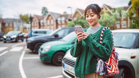 A young Asian woman making payment for parking place on mobile app with smartphone in car park.