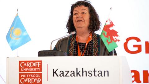 Menyw gyda gwallt brown yn sefyll tu ôl i ddarllenfa sy'n dweud Prifysgol Caerdydd Kazakhstan a baneri Kazakhstan a Chymru arno