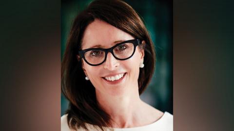 A shot of Cara looking straight into the camera, she has long dark hair, is wearing pearl-type earrings, a white top and glasses.