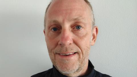 A balding man in a black top staring at the camera, he has light stubble that is mostly grey with flecks of black and the top of a black top is visible on his shoulders