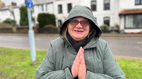 A blonde woman wearing a green waterproof coat, holding her hands together as if in prayer and looking straight at the camera. There's a grass verge, a road and a row of white houses in soft focus behind her.