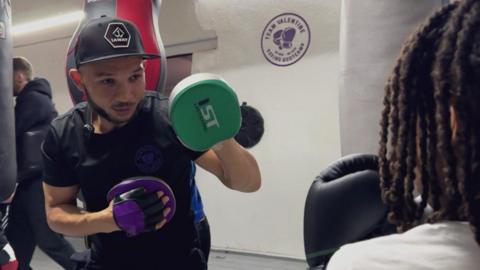 Curtis is wearing a snapback, he has boxing gloves on, and is in his gym. He is wearing a black t-shirt and looks focused.