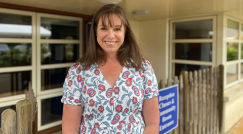 Fiona Vacher in a white dress with red and blue floral patterns stands in front of a building with large windows and a wooden fence. A blue sign in the background reads 'Reception, Classes & Community Room Entrance'.