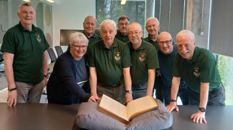 Sid Patterson, a trustee of the DLI Friends, centre, with Carolyn Ball, county archivist and manager of The Story, left, and Ian Brown, Chair of the DLI Friends, right, and other members of the Friends. They are smiling as the ledger rests on a big grey cushion in front of them. Sid Patterson has short white and is wearing a green t-shirt and grey trousers. Carolyn Ball has short white hair and blue long-sleeved top. She wears glasses and a necklace. Ian Brown has glasses and is wearing a blue top.