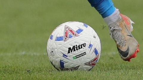 A National League football with HUFC written on it being kicked by a white boot with a blue sock 