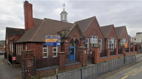Stirchley Primary School is a single-storey red brick building with pointed roofs and a blue door. A blue and yellow sign is outside the entrance. There are railings along the pavement and yellow road markings. The school windows are open.