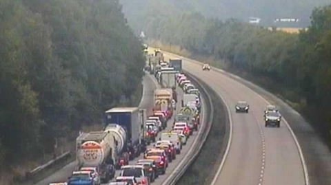 A motorway camera image of a motorway - there are two lanes of traffic queues on the left and a relatively empty carriageway - with just for cars visible - on the right. 