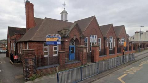 Stirchley Primary School is a single-storey red brick building with pointed roofs and a blue door. A blue and yellow sign is outside the entrance. There are railings along the pavement and yellow road markings. The school windows are open.