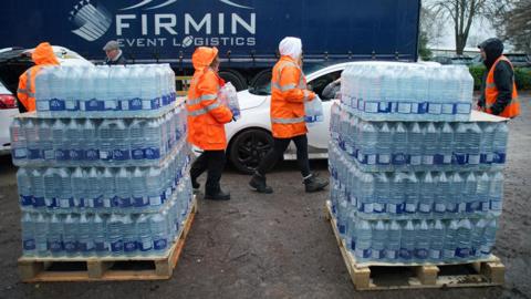 Two piles of bottled water sit on crates in front of a lorry. People in high vis orange jackets are delivering water to cars that are waiting
