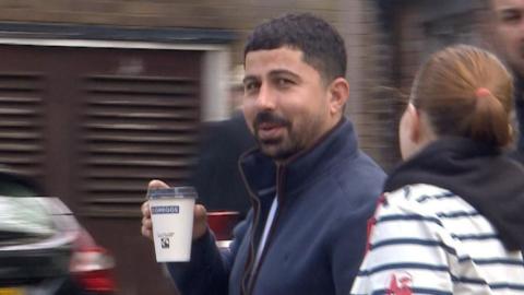 A man with short black hair and facial hair stares directly at the camera, mid-conversation with the woman next to him. He is wearing a navy zip-up hoodie and holding a coffee in a paper cup.