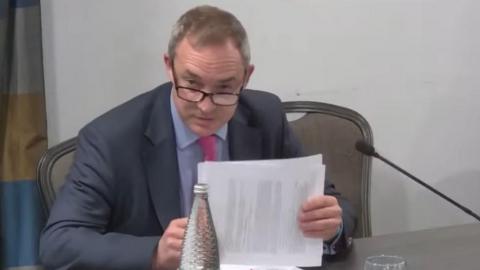 A still image of a livestream. Rob Sadler is speaking at the inquiry, holding papers in his hands. He has short grey hair and is wearing dark glasses, a navy suit, blue shirt and pink tie.