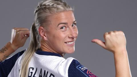 Bethany England in a blue and white England jersey. She has her hair in plaits and points at the back of her shirt.