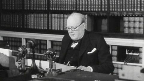 A black and white photo of Winston Churchill wearing glasses, sat behind a desk with microphones