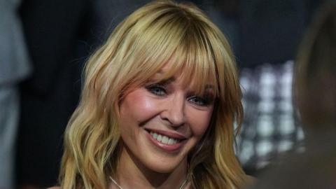 A close-up portrait of singer Kylie Minogue smiling. She has blonde, shoulder-length hair with a full fringe. She is wearing a pearl necklace. The background is blurred with dark tones and a grid pattern.