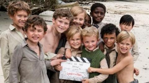 A group of 11 boys with a clipperboard on a beach.