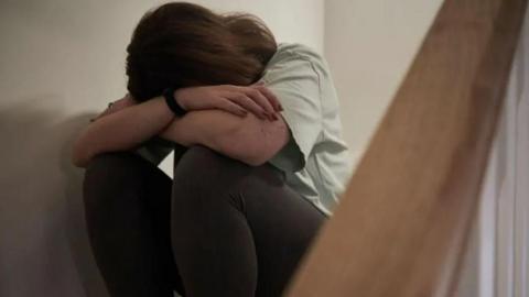 A woman sits on a staircase with her hand in her hands, she is wearing a white t-shirt and black trousers