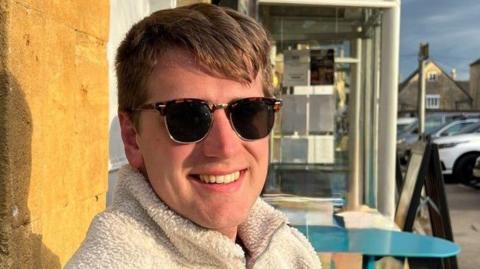A man with short brown hair and wearing sunglasses and a white fleece, smiles as he sits outside.