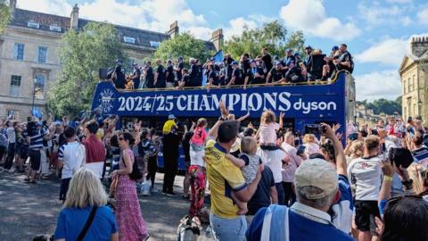Blue bus with 2024/2025 champions written on the side. Bath players are on the top of the bus dressed in black t shirts with blue Bath caps. Lots of people are waving and taking photos in the foreground.