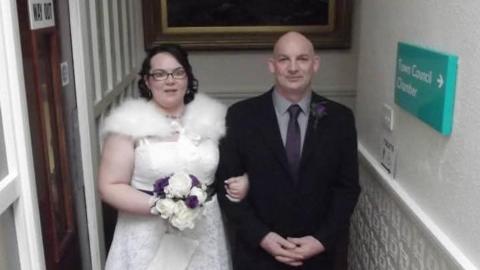 Brian Evans in a suit and tie next to his daughter Colette who is wearing a white wedding dress and is holding a bouquet of flowers. She is wearing glasses and has black hair. Mr Evans has a shaved head.