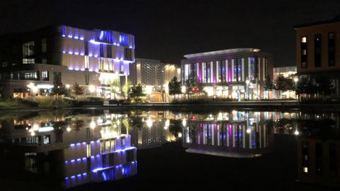 Several large buildings with multicoloured lights reflected in a large body of water in the foreground