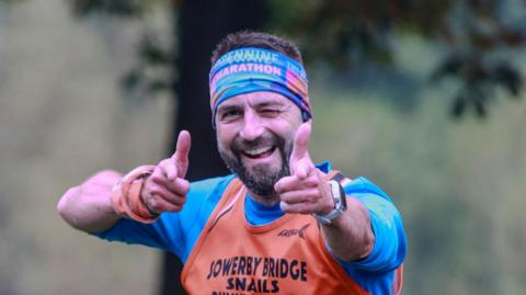 A man with a head band in bright colours wearing a blue T shirt with an orange vest over the top. He is smiling and pointing his fingers at the camera