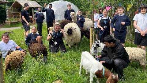 Pupils and teachers sit on a patch of grass with goats and sheep