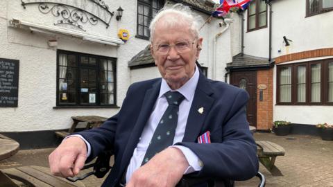 George Lewis sits in a walking aid outside a pub. He has white hair and wears a hearing aid and glasses. He wears a navy suit and dark patterned tie, and the ribbons of his two medals are visible behind his arm. 