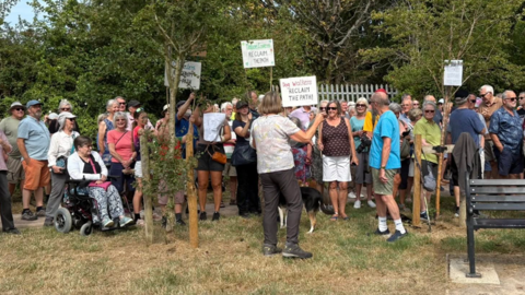 A group of protestors stand on the grass by trees. They are being spoken to by an organiser. Some protestors hold signs that say 'Dog walkers reclaim the path'.