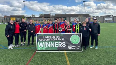 A line-up of players and coaching staff standing behind a placard which reads "Lincolnshire FA County Cup Final Winners".