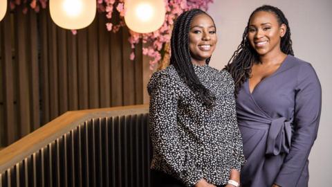 Two black women with long hair stand next to each other and are smiling at the camera while looking at it. One is wearing a dotty top and the other is wearing a purple dress. To the left of them is a bannister, lights hanging from the ceiling and pink flowers. 