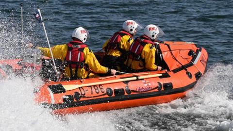 Three lifeboat crew in yellow coats, red lifevests and white helmets sporting "RNLI" in large red letters on board a small orange lifeboat motoring through the sea