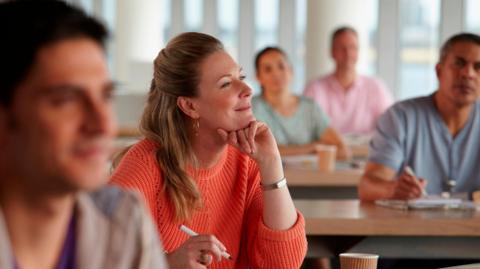 A stock image of a group of mature learners in a classroom. A blonde long-haired woman in an orange jumper is in focus, her chin resting on one hand and a pen in the other. She is smiling as she looks to the front. Three men and a woman sit on surrounding tables, out of focus.